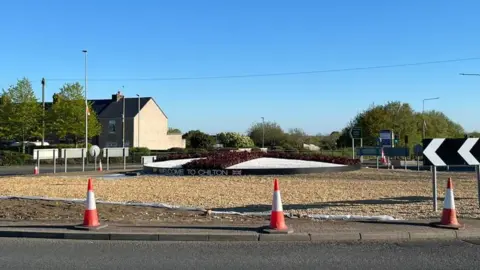 Golden brown gravel surrounds a raised bed reading 'Welcome to Chilton' on a roundabout. The roundabout has red plants and white gravel in the pattern of the St George's flag.