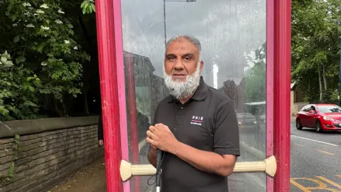 Sofía Luis-Hobbs/BBC Man wearing black short sleeve polo shirt stands in front of a red bus stop. He holds a cane in front of him as he looks into the camera to have his picture taken.
