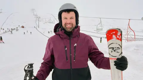 BBC News Skier Steve Fraser wearing a helmet and burgundy jacket stands on a snowy slope holding a ski, with chairlifts and other skiers visible in the background.