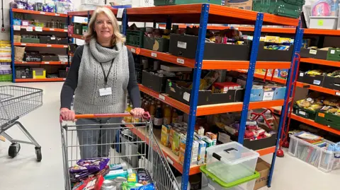 A women is in a foodbank, she is pushing a shopping trolley. 