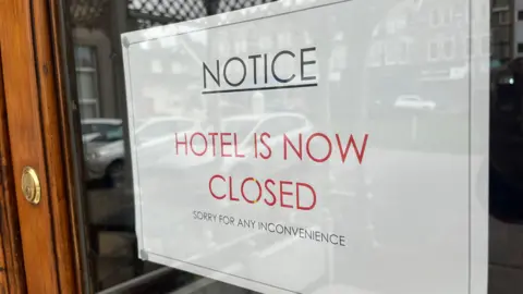 A sign placed in the glass window of a door reads Hotel is now closed, sorry for any inconvenience 