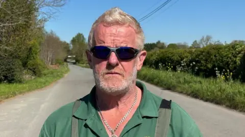Charlie Stubbs / BBC A man with short white hair and a white short beard. He is wearing black sunglasses with a slight dark blue tint. He is wearing a dark green polo shirt, which is unbuttoned, and a metal chain around his neck. He is standing the middle of a country road, which is surrounded by hedgerows