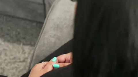 A woman is seen from behind and above. She has long dark hair, is sitting on a grey sofa and holds her hands together on her lap. Her nails are painted a bright turquoise.