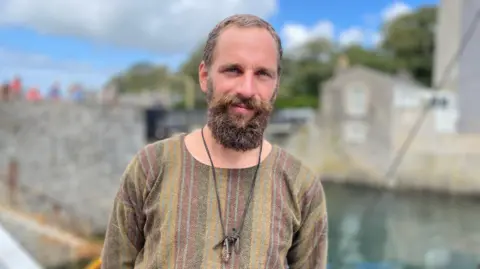 Ingwenog has short dark hair and a full beard. He's wearing a traditional woollen tunic with a leather necklace. He is standing in front of Castletown harbour, with stone walls, on a bright day.
