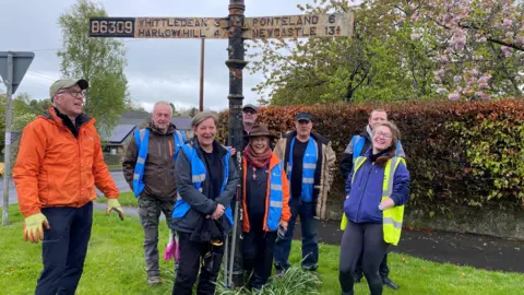A group of people all wearing brightly coloured hi-vis jackets and waterproofs are standing around a fingerpost which is very rusty and worn. The sign points in two directions. To the left, it indicates the B6309 and informs people that Whittledean is three miles away, Harlow Hill is four. To the right, Ponteland is six miles and Newcastle a very specific 13 and three quarters.