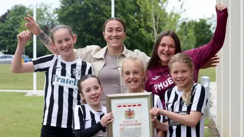 Lucy Bronze smiling and standing with five young girls in black and white football strips. She's got brown hair which is tied back and holding her arms out in celebration. The girls are holding a framed document which reads 'County of Northumberland'.