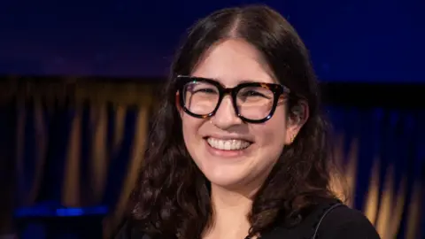 A woman wearing thick-rimmed dark glasses with long curly hair smiles. 