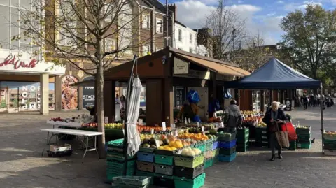 Alex Pope/BBC A market stall selling vegetables and fruit in a market square. Melons, oranges and other produce can be seen in crates at the front of the stall. A woman is walking away from the stall with two shopping bags and a man is talking to the stallholder. 