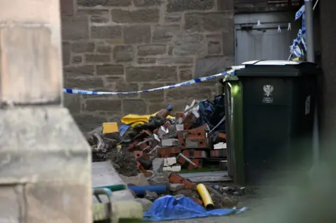 Stuart Cowper A pile of bricks and rubble on the ground with bins and police tape in the foreground