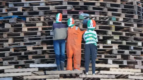 PA Media Effigies of three figures wearing balaclavas in the style of the green, white and orange Irish tricolour flag. The figure on the left is wearing a beige fleece and blue jeans, the one in the middle an orang boiler suit and the one on the right a Celtic top of green and white hoops and dark blue tracksuit bottoms. They are standing on the side of a bonfire made up of wooden pallets.
