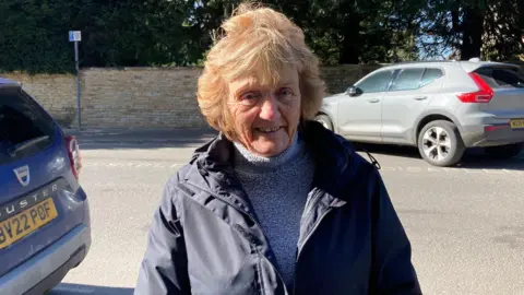 Janet Kirk. She is pictured outside. She has short fair hair and is wearing a grey jumper and a navy blue coat. She is looking directly at the camera and smiling.