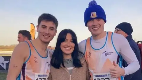 Jordan Adams Cian Adams and Jordan Adams are both wearing orange and white tank tops. Agnès Dangreaux is standing in the middle of them both, she has dark hair and is wearing a brown jumper
