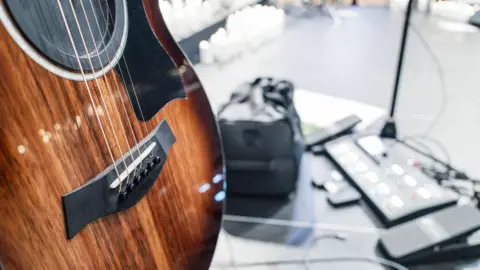Wooden acoustic guitar on its stand on a stage during rehearsals at a gig. Electronic amp floor pedals can be seen on the stage floor.