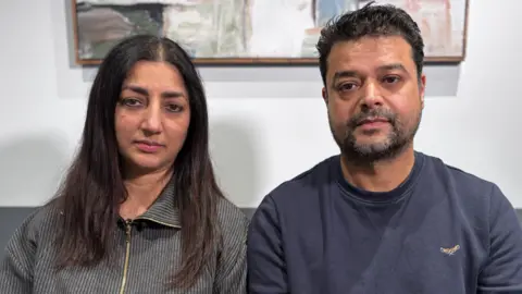 Aryan's parents Sukhi and Rakesh Sharma sitting side by side and looking at the camera with a painting in the background.