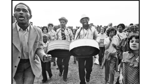 Diane Bush A black and white image of two steel drum players surrounded by children. 