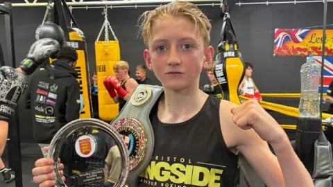 Archie with his winning belts and shield.  He has short blond hair and is wearing a black t-shirt with Bristol Ringside in yellow lettering.