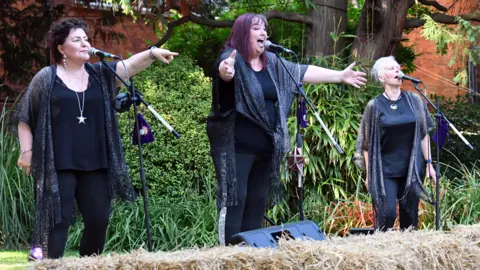 Jean Finch Ruth, Penny and Anita, singing in a garden, into three microphones. They are all wearing black outfits. Ruth, right is pointing one arm, Penny, in the middle has both arms out stretched and Anita on the right has her eyes closed and is singing into a microphone. There is a straw bale in front of them and an a speaker. 