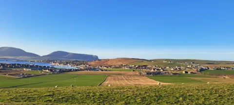 Duncan MacRae A view over the Orkney countryside with green fields and ploughed areas surrounding houses gathered round a bay with hills in the background and striking blue sky