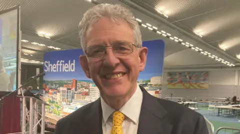 Simon Thake/BBC A man stands in a leisure centre where tables are in the background for the vote count. He has a black suit, yellow tie, wire frame glasses and grey hair. He is smiling widely.