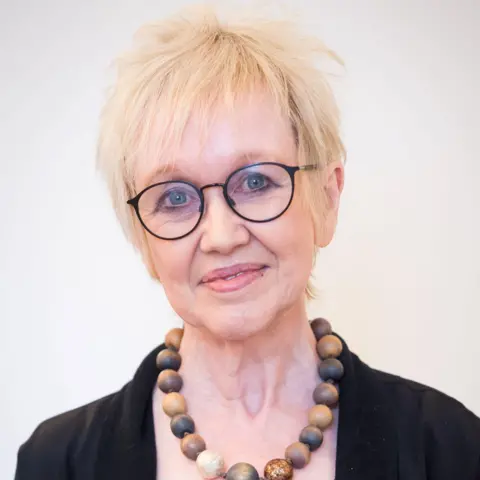 Royal College of Psychiatrists in Scotland Dr Jane Morris looking straight at the camera in front of a plain white background. She is wearing black framed glasses, a black jacket and brown wooden beads.