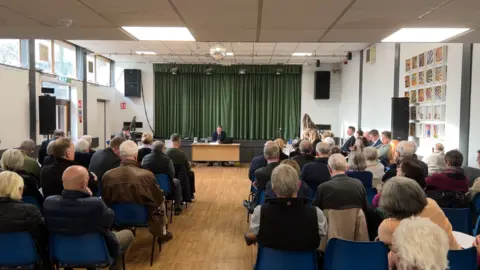 About 50 people sitting on blue plastic chairs in a village hall. They have their backs to the camera and are looking at a man at the front of the hall who is sitting behind a wooden table. There is stage above him with a green curtain across the front. 