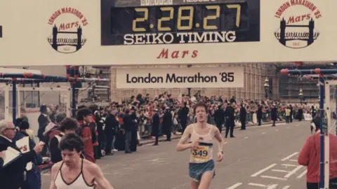 Chris at the finish line with a clock that says 2 hours and 28 min.