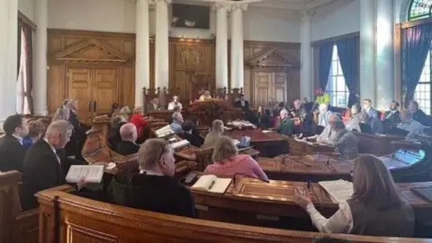 Councillors sat in a meeting inside South Shields Town Hall. They are sat at wooden benches in a semi-circle around the council leader, who is sat in a large chair at the front of the room.