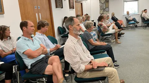 A photograph of people in the public viewing gallery within Cotswold District Councils chamber. There's nearly 2 dozen people siting on dark green chairs. They're various genders and ages generally wearing summer clothes. behind them is a white wall