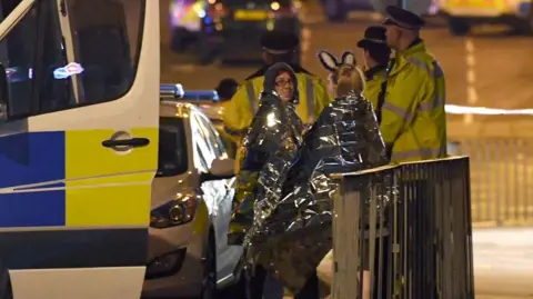 Getty Images Concert goers wrapped up in silver foil survival blankets wait to be picked up at the scene outside the Manchester Arena  