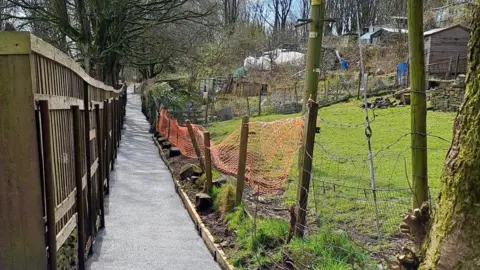 Bradford Council A tarmacked footpath with a fence on one side and green fields on the other. It is a sunny day in a semi-rural area