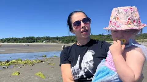 RTÉ A woman with long brown hair and pink shaded sunglasses, holds her child. The child has a pink floral bucket hat on, pink dress and sucks a yellow dummy. Her mother wears a black t-shirt with two full ringed hands on it. Behind her is the blue sea and green moss rocks that sit on the sand of the beach. People are walking along the sand in the background.