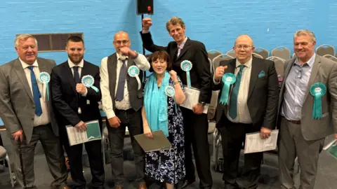 Seven Reform UK politicians are seen smiling and celebrating. They are wearing light blue rosettes and are stood against a blue wall.