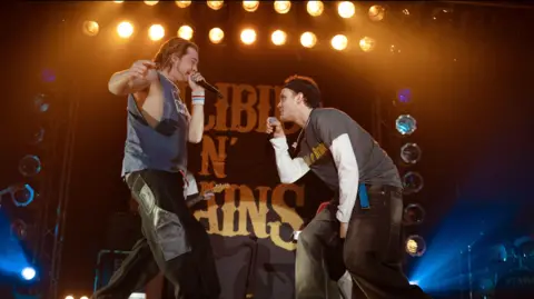 Mark Mainz Two men stand rapping onstage while looking at each other. They are wearing jeans and T-shirts.