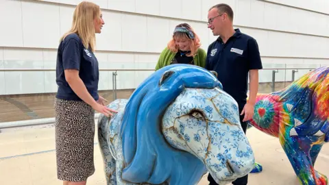 Sam looks down at her lion with two other people wearing branded tops. Behind them is a brightly coloured lion covered with a tie-dye print.