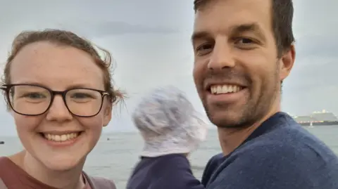 A smiling couple stand by the sea taking a selfie, with a baby in the man’s arms whose face is blurred by motion; a large ship is visible in the distance under a grey sky