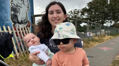 Ciara Harnden - a woman with dark hair holding a young baby and small toddler wearing sunglasses and a bucket hat.