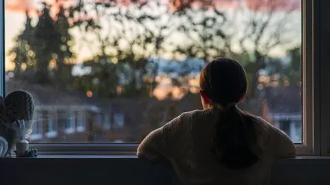 A young girl, with a ponytail, looks out of her bedroom window as the sun is setting. The view through the window is blurry but shows a residential area.
