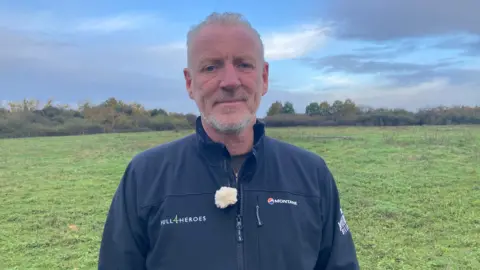 Jo Makel/BBC Paul Matson, who has short grey hair and a short grey beard, stands in a grassy field. He is wearing a blue jacket with the Hull 4 Heroes logo on.