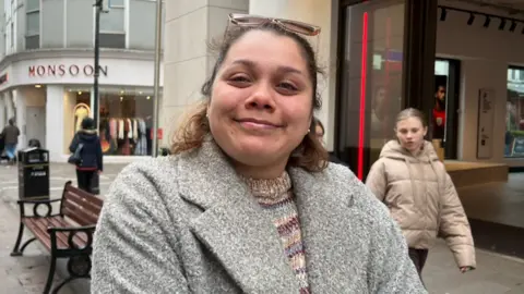 A woman standing on a busy shopping street near the entrance of a Sports Direct store. She's wearing a grey textured coat and a multicoloured knitted top, with a pair of sunglasses resting on her head.