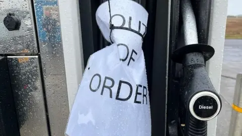 A close up of fuel pumps. The petrol nozzle has a paper sign wrapped round it that says "out of order".