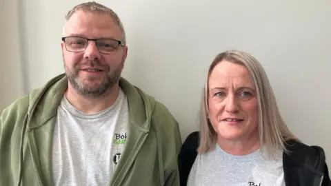 A man and woman are looking at the camera. Both wear grey t-shirts under their tops. The man has glasses and a short beard, the woman has shoulder length blonde hair