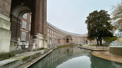 City Hall in Bristol. It is a grand, curved building with neatly lined windows and big pillars. There is water outside of it with a small fountain visible in the corner of the image.