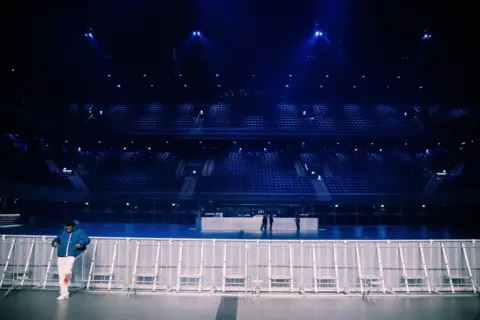 Oliver Akinfeleye Wizkid in a zipped-up blue puffer jacket, white trousers and white shoes leans against audience railings to the left of the shot in the Ziggo Dome in Amsterdam before a show in November 2022.
