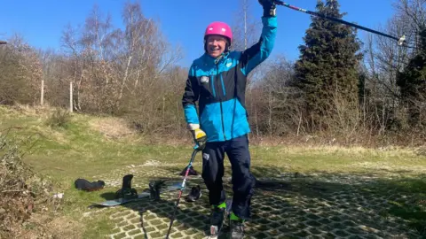 BBC/Simon Thake A man in a ski jacket, trousers and helmet stands in skis at the top of a worn dry ski slope.