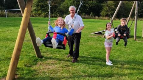 An older man playing pushing a girl on the swing with other children playing nearby