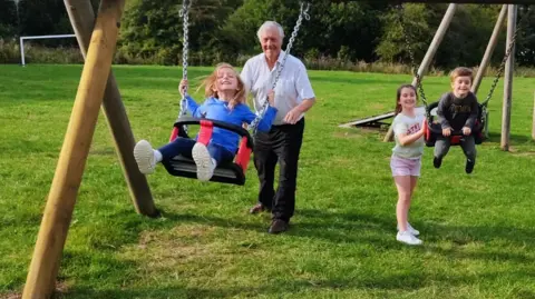 LDRS An older man playing pushing a girl on the swing with other children playing nearby