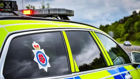 A police car is parked facing a road. It has a sticker on the back window which reads Lancashire Constabulary. A blue sky and some trees can be seen in the distance over a road.