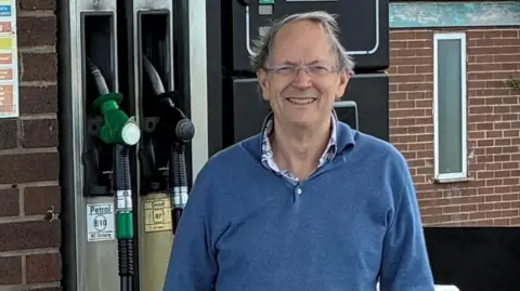 Peter Nicholson has short grey hair and is wearing glasses and a blue jumper