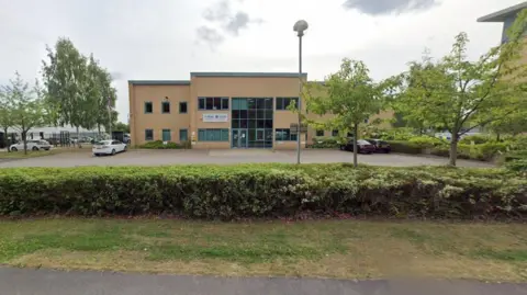 Google A Street View image of a two-storey building. A car park is in front of the building with three cars parked. To the left of the central glass front is a sign with the force logo. Hedges and trees surround the car park.