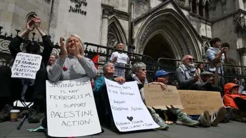 About a dozen Palestine Action supporters are seen outside of the Royal Courts of Justice on Monday morning. Six placards in support of the organisation are seen being held by people 